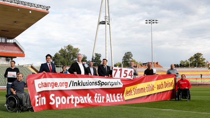 Eine Gruppe von Menschen, darunter 2 Rollstuhlnutzende, hält ein riesiges Transparent mit der Aufschrift "Ein Sportpark für ALLE!". Sie stehen dabei auf dem Rasen eines Stadions.